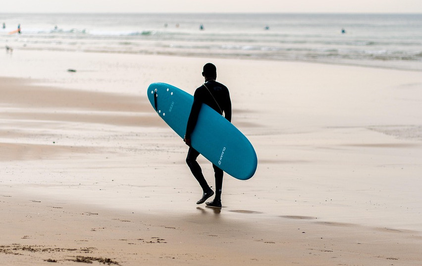 A surfer is walking along the shoreline, holding his surfboard while checking the wave conditions at Kuta Beach, Bali.