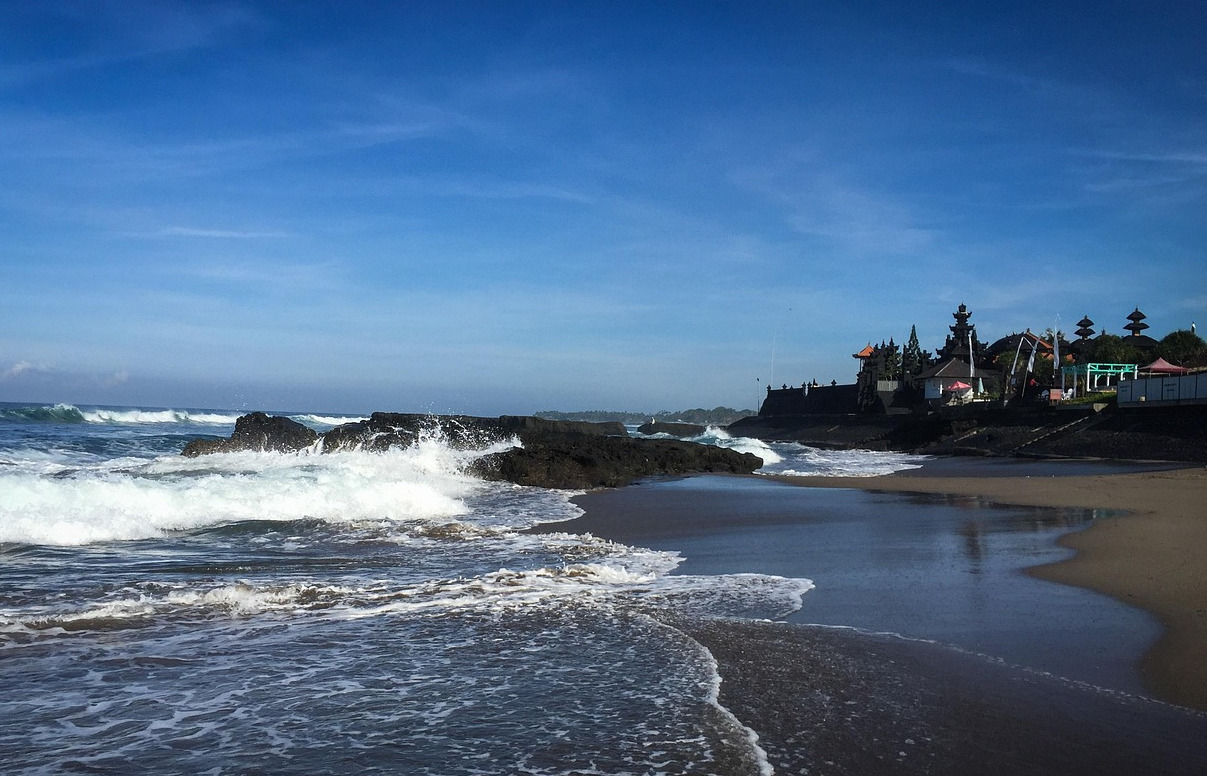 The afternoon atmosphere at Batu Bolong Beach in Pererenan, Canggu, featuring waves, rocky outcrops, and exotic black sand, set against the backdrop of a temple.