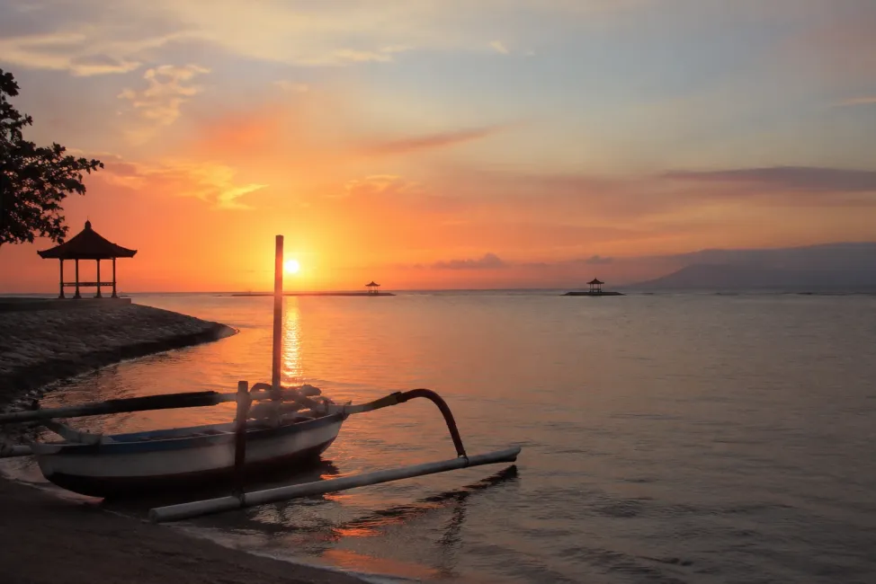 A wide shot of Sanur's calm beach at sunrise, soft pastel sky reflected on gentle water, a traditional outrigger boat silhouette